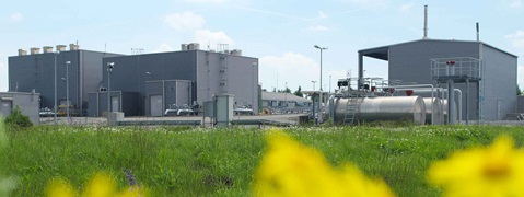 Industrial buildings with silver exteriors and pipes, set against a green field with wildflowers under a blue sky.